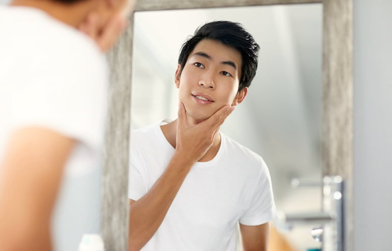 Young man who eats collagen-rich foods with glowing, youthful skin looking at his complexion in the mirror